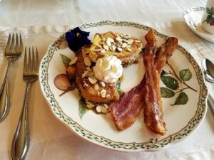 Creme Brulee French toast on china plate with bacon over set table. 