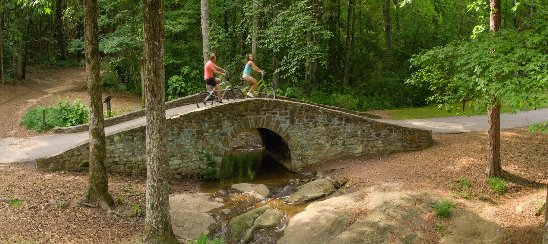 A man and woman on bikes riding over a stone bride in wooded area.