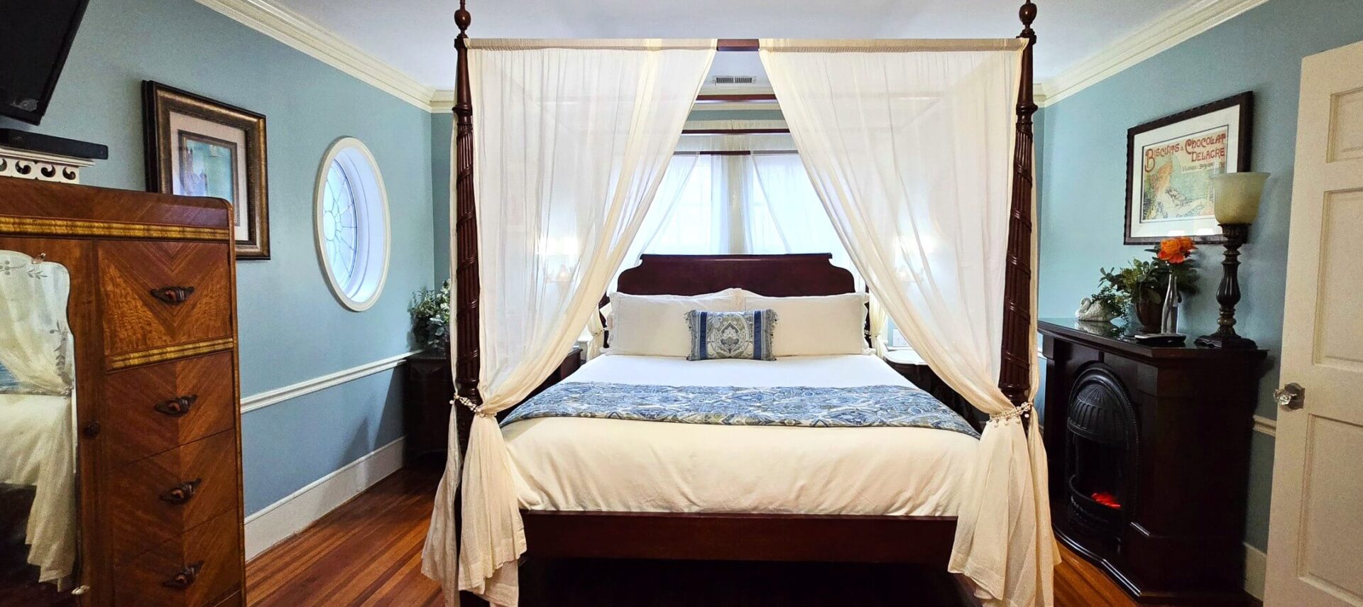 California King bed with four posters and curtains near electric fireplace.