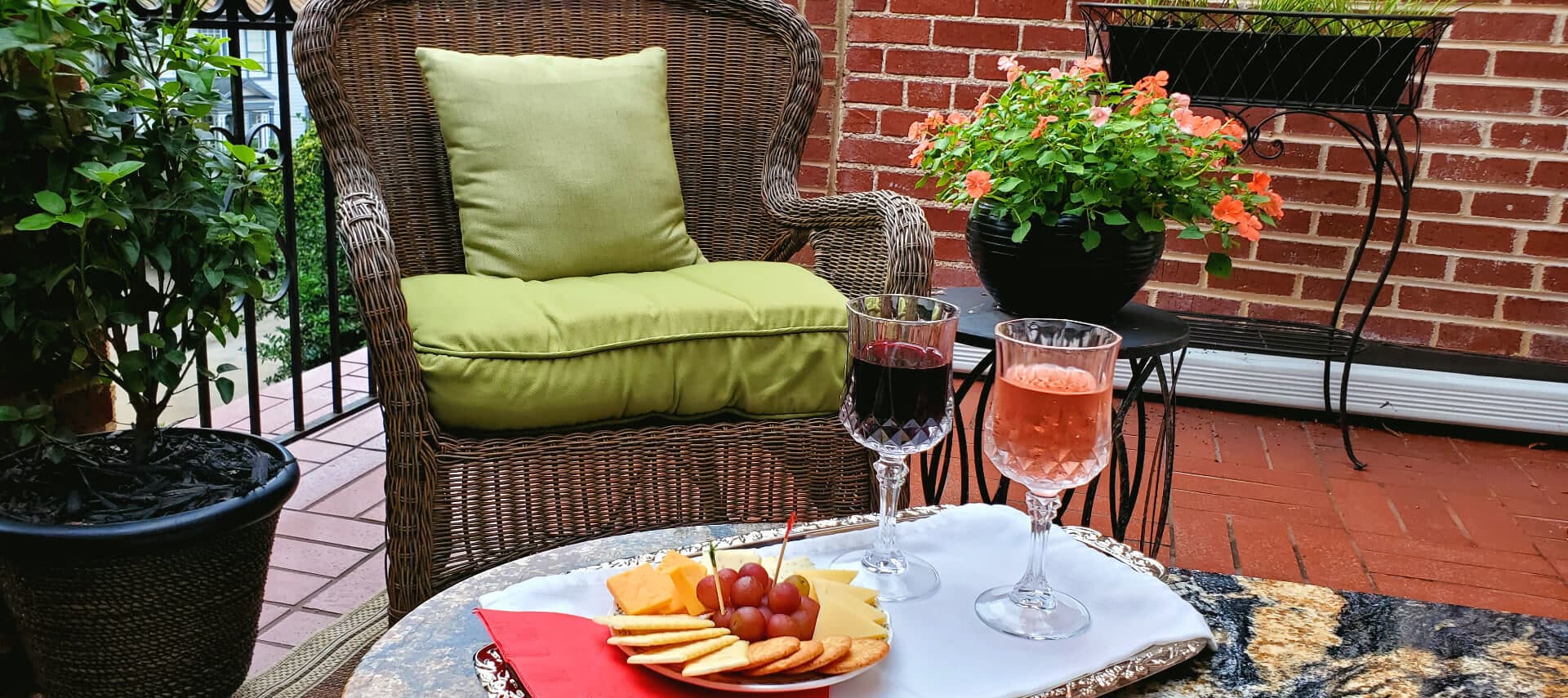 Wine, cheese, and crackers served on silver tray over marble table on brick balcony with flowers and chair.